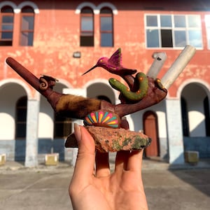 May include: A hand-painted ceramic pipe with a hummingbird, snake, and crystal accents. The pipe is resting on a rock with a painted design.