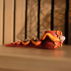 May include: A red and orange ceramic pipe shaped like a snake with a swirl design on the top. The pipe is decorated with a small, white, iridescent stone.