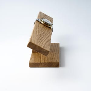 May include: A wooden watch stand with a watch resting on the top. The stand is made of light brown wood and has a simple design.