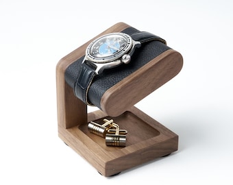 May include: A wooden watch stand with a dark brown grain finish features a black leather-textured cushion holding a silver-toned wristwatch with a blue face. A small square tray at the base contains a pair of gold and silver-toned metal cufflinks.