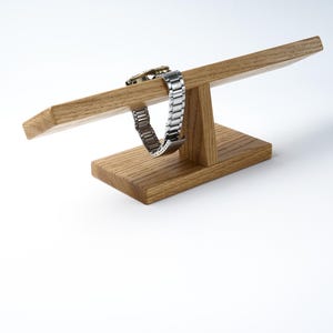 May include: A wooden watch stand with a watch band resting on the top. The stand is made of light brown wood and has a simple design.