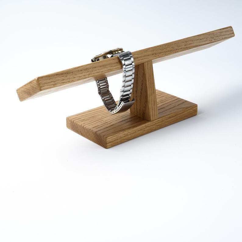May include: A wooden watch stand with a watch band resting on the top. The stand is made of light brown wood and has a simple design.