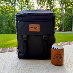 May include: Black insulated backpack cooler with a brown leather patch that says "DADDY". A brown leather can cooler with the text "WE LOVE YOU GEORGIA MILES" is next to the backpack. The background is a green, natural setting.