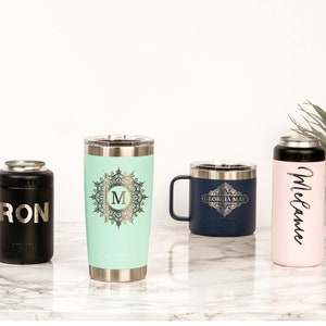 May include: A collection of personalized drinkware, including a black can cooler with "RON" printed on it, a mint green tumbler with a monogram, a navy blue mug with "GEORGIA MAY", and a pink can cooler with "Melanie" in script. All items are stainless steel.