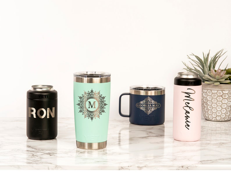 May include: A collection of personalized drinkware, including a black can cooler with "RON" printed on it, a mint green tumbler with a monogram, a navy blue mug with "GEORGIA MAY", and a pink can cooler with "Melanie" in script. All items are stainless steel.
