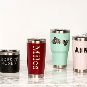 May include: Four personalized insulated tumblers and a mug with names engraved. The tumblers are in black, red, mint green, and pink. The mug is black. The names are "Robert Jones", "Miles", "Stacy", and "Ann".