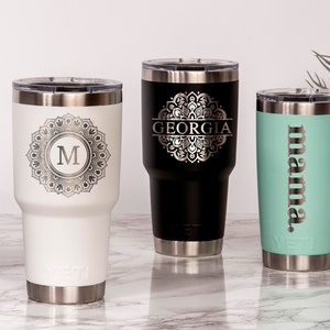 May include: A collection of personalized stainless steel tumblers in various colors. One white tumbler features a mandala design with the letter "M", a black tumbler has the name "GEORGIA", and a teal tumbler says "mama".