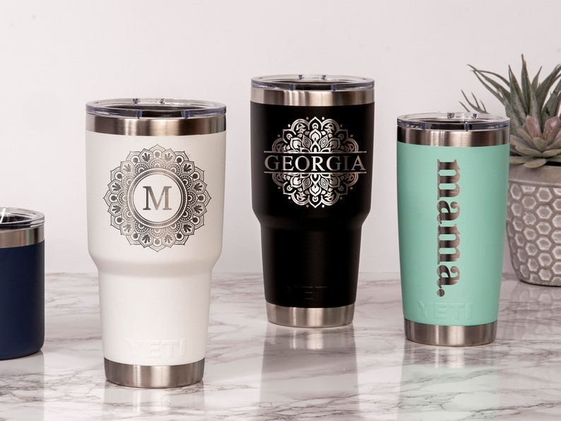 May include: A collection of personalized stainless steel tumblers in various colors. One white tumbler features a mandala design with the letter "M", a black tumbler has the name "GEORGIA", and a teal tumbler says "mama".
