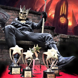 Pode incluir: Uma pessoa vestida como um rei esqueleto com uma jaqueta e calças de couro preto senta-se em um trono de tijolo segurando um troféu. A pessoa está usando uma coroa e segurando um cetro. O fundo é vermelho e laranja fogo com uma estrutura alta e escura à distância. A pessoa está cercada por cinco troféus dourados em forma de estrela.