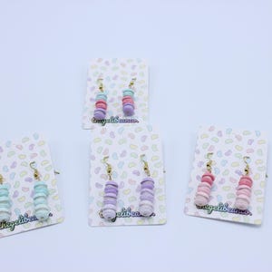 May include: Three pairs of pastel-colored dangle earrings on white card stock with a jelly bean pattern. The earrings are made of polymer clay and feature a stack of small circles in various shades of pink, purple, and blue.