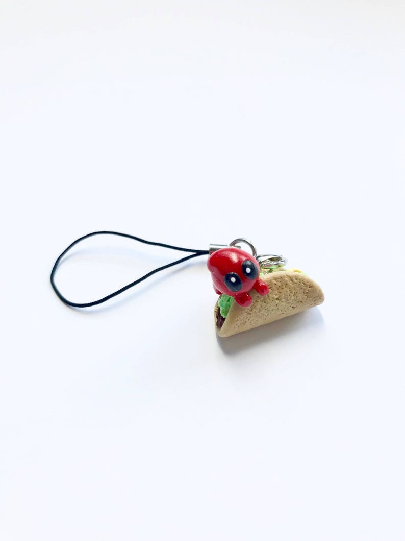 May include: A red, cartoon-like character with black eyes and a black string attached to a miniature taco charm. The taco has a light brown shell, green lettuce, and a small amount of filling. The charm is on a white background.