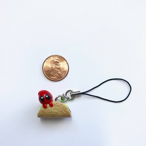 May include: A small, handmade charm featuring a red character with black eyes perched on a miniature taco. The taco is tan with green lettuce and a silver clasp with a black cord. A penny is next to the charm.