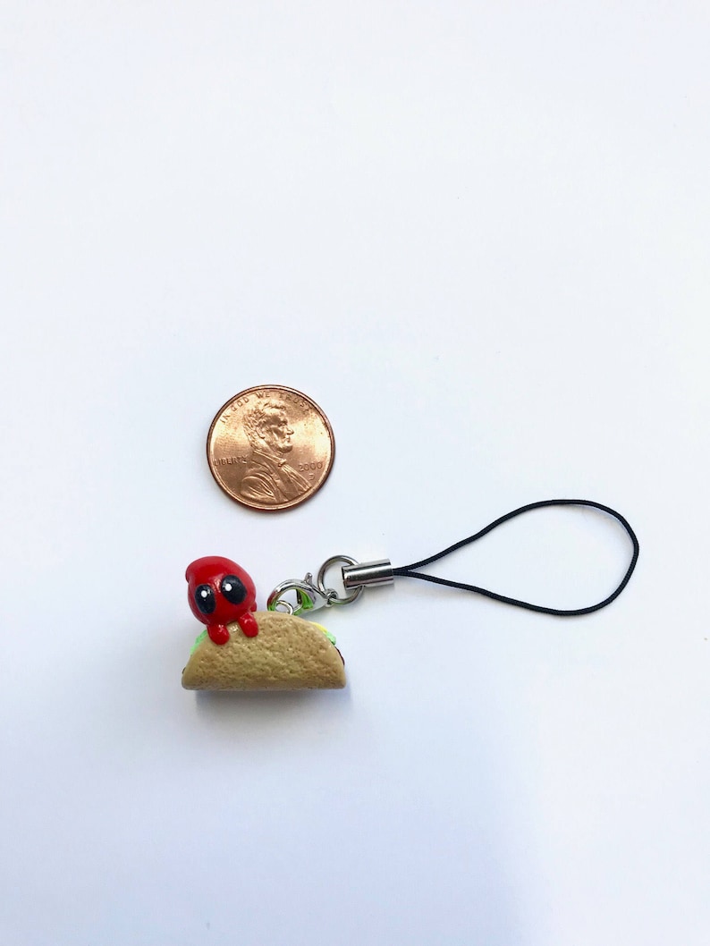 May include: A small, handmade charm featuring a red character with black eyes perched on a miniature taco. The taco is tan with green lettuce and a silver clasp with a black cord. A penny is next to the charm.