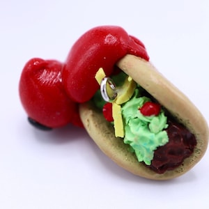 May include: A whimsical, handmade taco sculpture with a red boxing glove holding the taco. The taco features a light brown shell, green guacamole, red tomatoes, yellow cheese, and a silver ring. The sculpture is set against a white background.