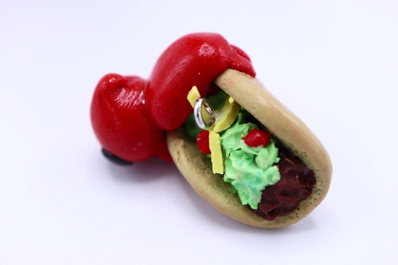 May include: A whimsical, handmade taco sculpture with a red boxing glove holding the taco. The taco features a light brown shell, green guacamole, red tomatoes, yellow cheese, and a silver ring. The sculpture is set against a white background.