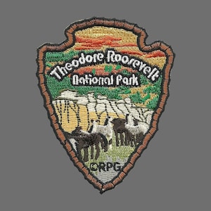 May include: A brown, green, and yellow embroidered patch with the text "Theodore Roosevelt National Park" and a depiction of bison in a grassy field. The patch is shaped like a teardrop and has a brown border.