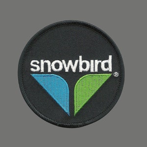 Peut inclure: Un écusson brodé noir avec le mot "snowbird" en lettres blanches et un motif d'oiseau stylisé vert et bleu.