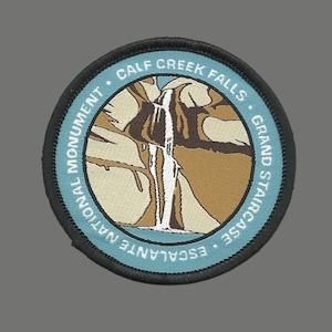 May include: A round patch featuring a waterfall scene with the text "CALF CREEK FALLS" and "GRAND STAIRCASE ESCALANTE NATIONAL MONUMENT" in a circular design. The patch has a blue border and a black stitched edge.