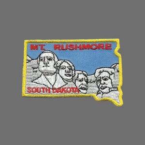 May include: A yellow embroidered patch with the text "MT. RUSHMORE" and "SOUTH DAKOTA" in red. The patch depicts a white outline of Mount Rushmore with the four presidents carved into the mountain.