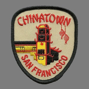 May include: A beige embroidered patch with a black outline. The patch features a red embroidered image of a pagoda with a yellow roof and red lanterns hanging from the top. The text "CHINATOWN SAN FRANCISCO" is embroidered in red.