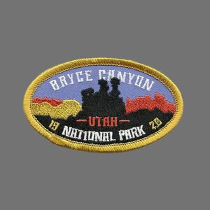 May include: A yellow and gold oval embroidered patch with the text "Bryce Canyon National Park" and "Utah" in white. The patch features a silhouette of a rock formation with a red and orange sunset behind it.