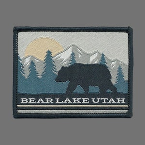 Peut inclure: Un patch brodé gris et bleu avec une silhouette d'un ours marchant devant une chaîne de montagnes avec un soleil jaune en arrière-plan. Le texte "BEAR LAKE UTAH" est brodé en blanc au bas du patch.