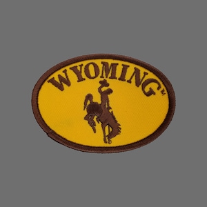 May include: A yellow oval patch with a brown embroidered outline. The patch features the word "WYOMING" in brown lettering and a brown silhouette of a bucking bronco.