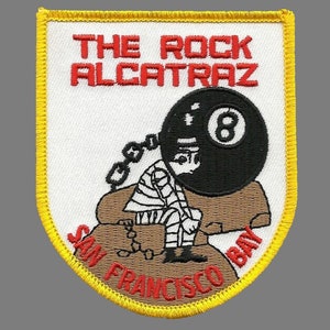 May include: A yellow and white embroidered patch with the text "The Rock Alcatraz San Francisco Bay" and a cartoon image of a person in chains holding a black 8-ball.