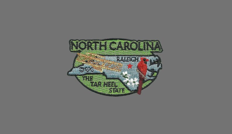 May include: Embroidered patch featuring a green outline of North Carolina with a red cardinal bird, the text "North Carolina", "The Tar Heel State", and "Raleigh" inside the outline.