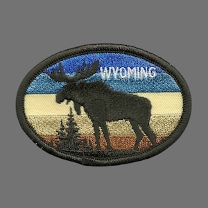 May include: An oval-shaped embroidered patch featuring a black silhouette of a moose standing in front of a multi-coloured sunset. The text "WYOMING" is embroidered in white on the blue part of the sunset.