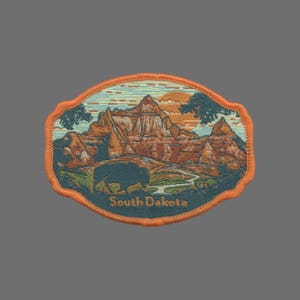 May include: An orange embroidered patch with a sunset scene of a mountain range and a buffalo in the foreground. The text "South Dakota" is embroidered in orange at the bottom of the patch.