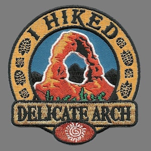 May include: A round, embroidered patch with a yellow border and the text "I HIKED DELICATE ARCH" in black. The center of the patch features a colorful image of Delicate Arch in Arches National Park, Utah.