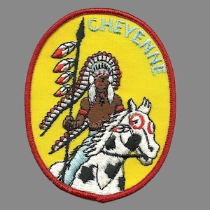 May include: A yellow embroidered patch with a red border. The patch features a Native American man riding a white horse. The man is wearing a feathered headdress and holding a spear. The text "CHEYENNE" is written in a blue and white font around the edge of the patch.