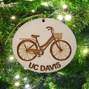 May include: A round wooden ornament with a laser-etched design of a bicycle with a basket. The ornament is decorated with the text "UC DAVIS".