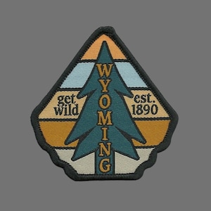 May include: A brown, blue, and white embroidered patch with a pine tree design. The text "WYOMING" is vertically centered in the tree, with "get wild" on the left and "est. 1890" on the right.