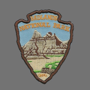 May include: A brown embroidered patch with a blue background and the text "Badlands National Park". The patch features a stylized image of a mountain range.