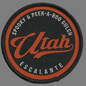 May include: A round, black patch with an orange border. The text "Spooky & Peek-A-Boo Gulch Utah Escalante" is written in white around the edge of the patch. The word "Utah" is written in orange in a large, cursive font in the center of the patch.