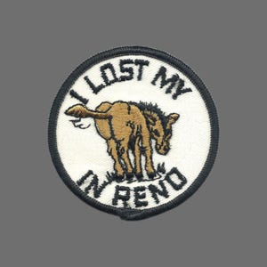 May include: Embroidered patch with a white background and a black border. The patch features a brown donkey with its rear end facing the viewer, standing on green grass. The text "I LOST MY" is arched above the donkey, and "IN RENO" is arched below.