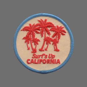 May include: A round, embroidered patch with a light beige background and a blue border. Three red palm trees are embroidered in the centre of the patch. The text "Surf's Up CALIFORNIA" is embroidered in red below the palm trees.