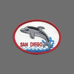 May include: Embroidered patch featuring a gray dolphin leaping over blue waves. The patch has a white background with a red border and the words "SAN DIEGO" in red. A souvenir item.
