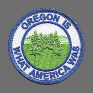 May include: Embroidered patch with a blue border and the text "OREGON IS WHAT AMERICA WAS". The patch features a green field with two green trees and a white background.