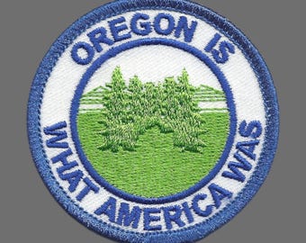May include: Embroidered patch with a blue border and the text "OREGON IS WHAT AMERICA WAS". The patch features a green field with two green trees and a white background.