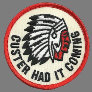 May include: A white circular patch with a red border. The patch features a black and white illustration of a Native American wearing a headdress. The text "Custer Had It Coming" is written in black around the illustration.