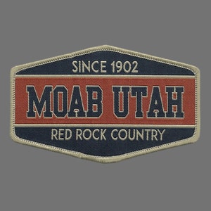 May include: A tan and blue embroidered patch with the text "Moab Utah" in navy blue lettering. The patch also includes the text "Since 1902" and "Red Rock Country".