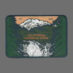 May include: Green embroidered patch with the text "Mt. Rainier National Park" in orange, "Est. 1899" in white, and a white mountain range illustration.