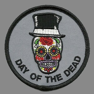 May include: A round, gray embroidered patch with a black and white sugar skull wearing a top hat. The skull is decorated with colorful flowers and the text "Day of the Dead" is written around the skull.