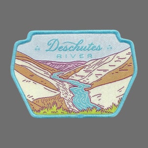 May include: Embroidered patch featuring a scenic landscape of the Deschutes River. The design includes a winding blue river through a canyon with tan, purple, and green accents. The text "Deschutes River" is embroidered in a light blue script.