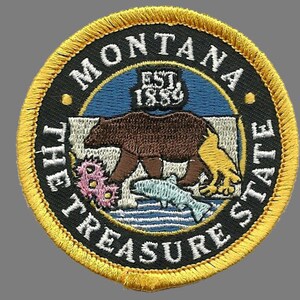 May include: A round, yellow-trimmed patch with the words "MONTANA THE TREASURE STATE" and "EST. 1889" in white text on a black background. The center of the patch features a brown bear, a yellow bird, and a blue fish on a blue and white background.