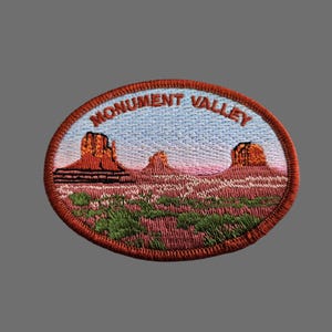 May include: Oval embroidered patch featuring a landscape of Monument Valley. The patch depicts red rock formations under a blue sky with the words "Monument Valley" arched across the top in brown thread. Green and pink details fill the foreground.