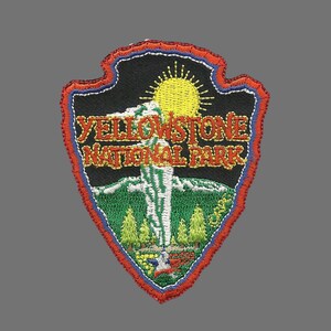 May include: Yellowstone National Park embroidered patch with a red, blue and white border. The patch features a black background with a yellow sun, a white geyser and green trees. The text "Yellowstone National Park" is in yellow and orange.